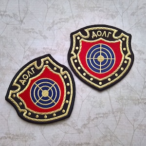 May include: Two black and gold embroidered patches with a red and blue target in the centre. The patches are shaped like shields and have the word "ДОЛГ" written in gold lettering at the top.
