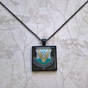 May include: A black chain necklace with a square pendant featuring a green, blue, and gold emblem. The emblem is a stylized eagle with wings spread and a shield in the center.