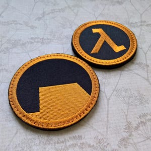 May include: Two circular embroidered patches featuring the Half-Life logo. Each patch has a black background with a golden yellow lambda symbol or a stylized golden yellow shape, both bordered by a thick golden yellow stitched edge.