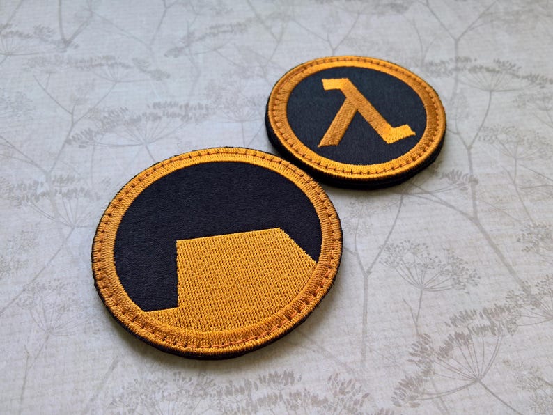 May include: Two circular embroidered patches featuring the Half-Life logo. Each patch has a black background with a golden yellow lambda symbol or a stylized golden yellow shape, both bordered by a thick golden yellow stitched edge.
