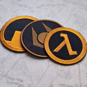 May include: Three circular embroidered patches featuring black fabric with gold-colored thread designs. The patches display distinct symbols, including a lambda character, a stylized geometric emblem, and a partial circular graphic with a gold border.