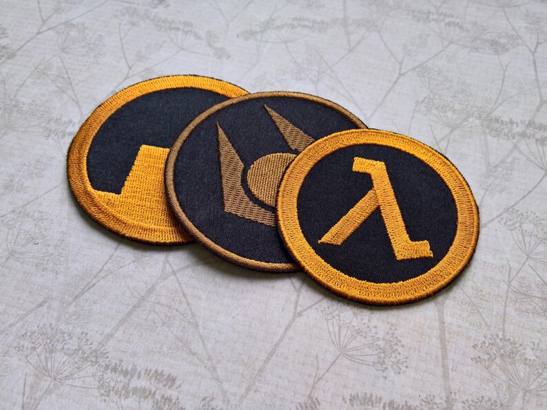 May include: Three circular embroidered patches featuring black fabric with gold-colored thread designs. The patches display distinct symbols, including a lambda character, a stylized geometric emblem, and a partial circular graphic with a gold border.