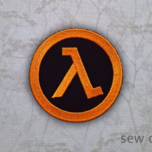 May include: Orange and black sew-on patch featuring the letter "A" in a stylised font. The patch is circular and has a black background with an orange border.