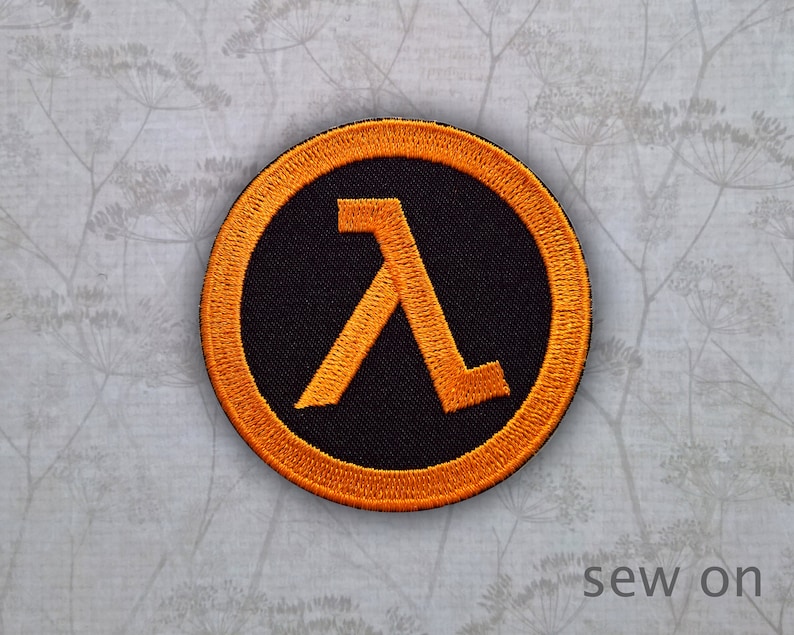 May include: Orange and black sew-on patch featuring the letter "A" in a stylised font. The patch is circular and has a black background with an orange border.