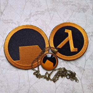 May include: Two circular embroidered patches featuring black and orange Half-Life logo designs, accompanied by a round pendant necklace with a matching orange and black graphic, suspended on a thin metallic chain with a lobster claw clasp.