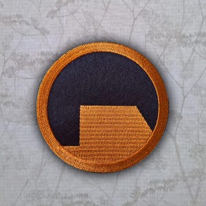 May include: A circular embroidered patch featuring a dark navy blue background with a stylized orange lambda symbol. The patch has a thick, textured orange border that frames the central design, creating a distinct contrast between the colors.