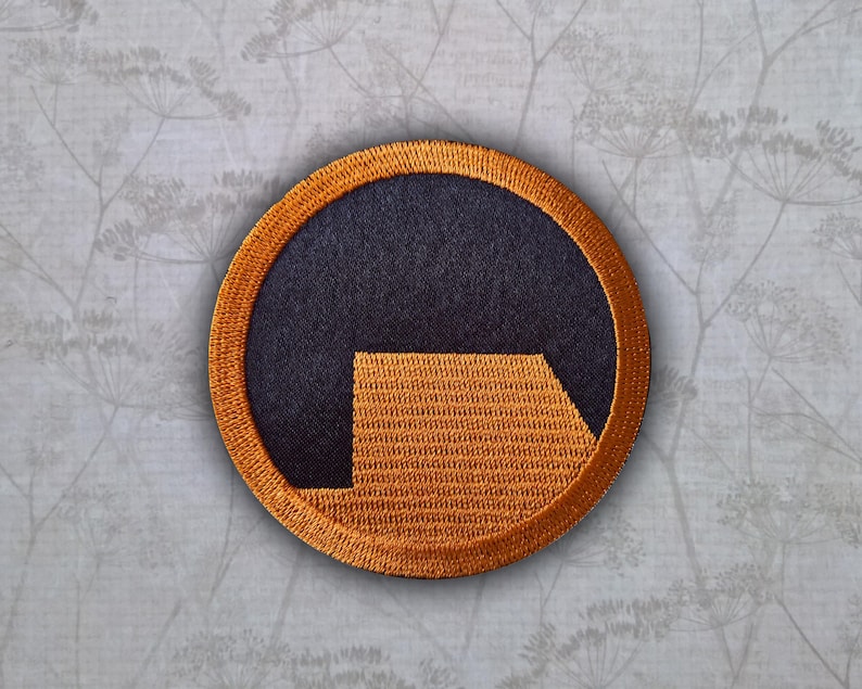 May include: A circular embroidered patch featuring a dark navy blue background with a stylized orange lambda symbol. The patch has a thick, textured orange border that frames the central design, creating a distinct contrast between the colors.