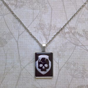 May include: A silver chain necklace with a black and white skull pendant. The pendant is in a silver frame and has a dark background.