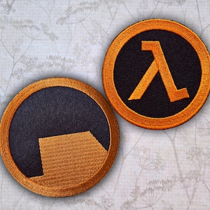 May include: Two circular embroidered patches featuring the Half-Life logo. One patch displays the orange Greek letter lambda on a black background, while the other shows a stylised orange geometric shape on a black background, both with orange borders.