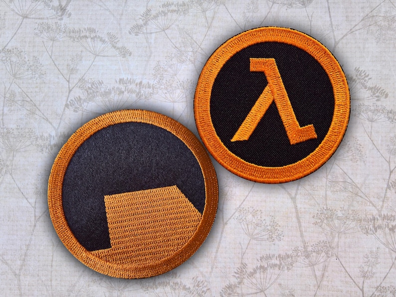 Pu&ograve; includere: Due toppe ricamate circolari con il logo di Half-Life. Una toppa mostra la lettera greca lambda arancione su sfondo nero, mentre l'altra mostra una forma geometrica arancione stilizzata su sfondo nero, entrambe con bordi arancioni.