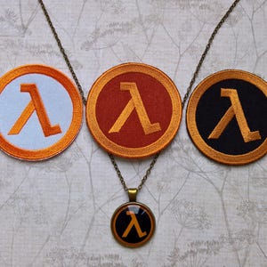 Op de afbeelding: Drie ronde geborduurde patches met de Griekse letter lambda in het oranje. Eén patch heeft een witte achtergrond, één een roestoranje achtergrond en één een zwarte achtergrond. Een bronskleurige hanger aan een ketting met een zwart lambda-icoon.