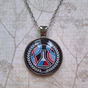 Peut inclure: Un collier en argent avec un pendentif rond représentant une illustration en noir et blanc d'un bécher contenant un liquide bleu et rouge. Le bécher est entouré d'une couronne de feuilles.