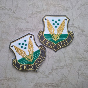 Puede incluir: Dos parches bordados con un diseño de escudo verde y blanco. El escudo presenta un águila dorada estilizada con las alas extendidas y la palabra "EKOLOG" escrita en un arco negro alrededor de la parte inferior.
