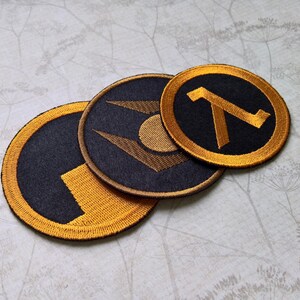May include: Three circular embroidered patches featuring black and gold thread. One patch displays a lambda symbol, another shows a geometric emblem with a central circle, and the third features a stylized block design with a gold border.