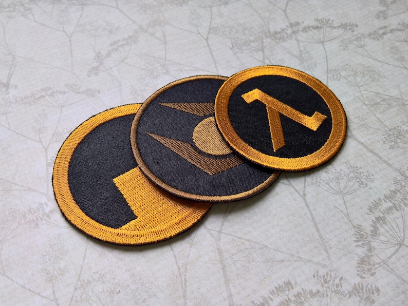 May include: Three circular embroidered patches featuring black and gold thread. One patch displays a lambda symbol, another shows a geometric emblem with a central circle, and the third features a stylized block design with a gold border.
