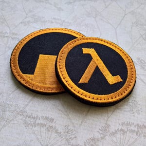 May include: Two circular embroidered patches featuring the orange and black Half-Life lambda symbol. The larger patch displays the lambda icon, while the smaller patch shows a partial view of the same design with orange stitching on black fabric.