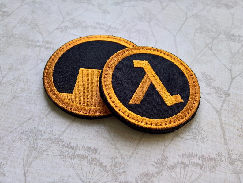 May include: Two circular embroidered patches featuring the orange and black Half-Life lambda symbol. The larger patch displays the lambda icon, while the smaller patch shows a partial view of the same design with orange stitching on black fabric.