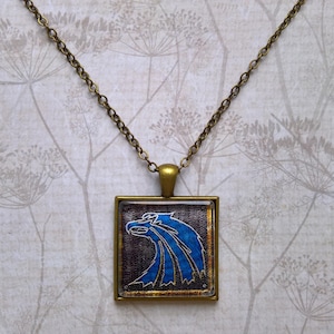 May include: A bronze chain necklace with a square pendant featuring a blue and gold eagle design.