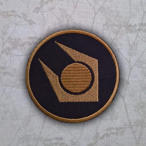 May include: A circular embroidered patch featuring a dark navy blue background with a gold-colored Lambda symbol centered on the fabric. The patch is finished with a gold-colored stitched border that frames the entire circular design.