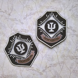 May include: Two black and white embroidered patches in a shield shape. Each patch features a white geometric design, a symbol, and text in a banner. The patches are set against a patterned background.