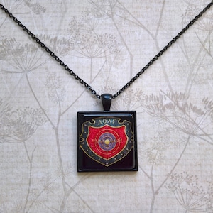 May include: A black chain necklace with a square pendant featuring a red and gold shield with the word "ROME" written on it.
