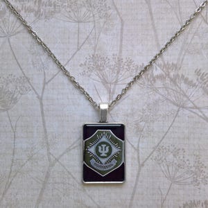 May include: Silver-toned necklace with a rectangular pendant. The pendant features a dark background with a green and white emblem. The emblem has a symbol and text in a foreign language. The chain is a delicate link style.
