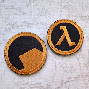 May include: Two circular embroidered patches featuring black and orange thread. One patch displays a solid orange geometric shape on a black background, while the other patch features the orange Greek letter lambda symbol centred on a black field.
