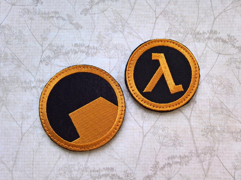 May include: Two circular embroidered patches featuring black and orange thread. One patch displays a solid orange geometric shape on a black background, while the other patch features the orange Greek letter lambda symbol centered on a black field.