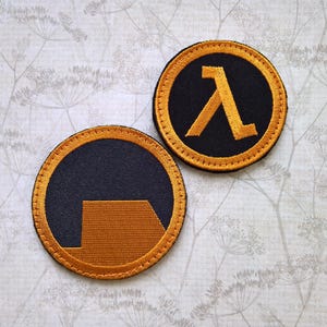 May include: Two circular embroidered patches featuring the Half-Life video game logo. One patch displays the Greek letter lambda in gold thread on a black background, while the other shows a gold geometric shape on a black background.