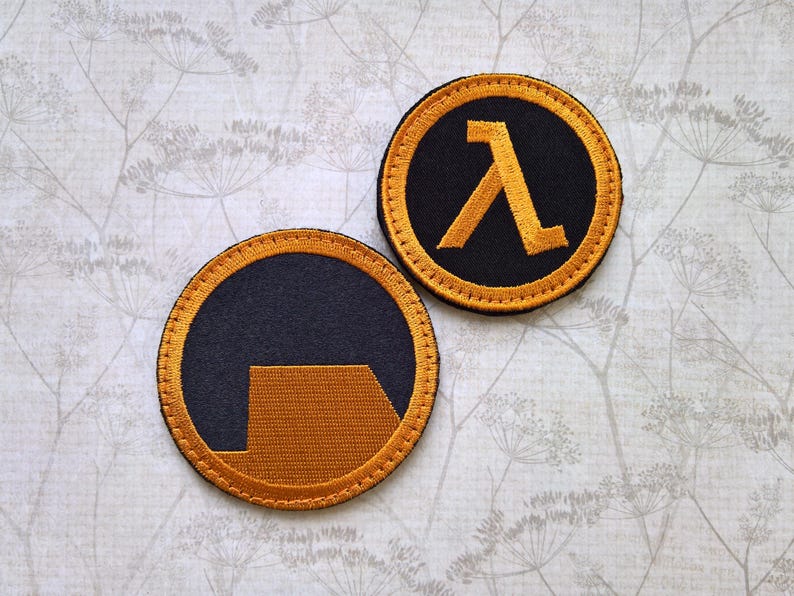 May include: Two circular embroidered patches featuring the Half-Life video game logo. One patch displays the Greek letter lambda in gold thread on a black background, while the other shows a gold geometric shape on a black background.