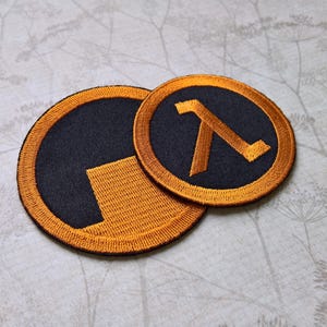 Pu&ograve; includere: Due toppe ricamate circolari con il design del simbolo Lambda in arancione e nero. La toppa pi&ugrave; grande mostra un blocco arancione parziale su sfondo nero, mentre quella pi&ugrave; piccola mostra la lettera greca lambda completa in arancione.