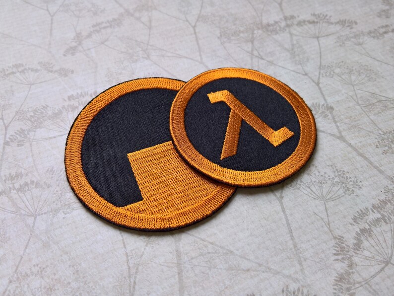 Pu&ograve; includere: Due toppe ricamate circolari con il design del simbolo Lambda in arancione e nero. La toppa pi&ugrave; grande mostra un blocco arancione parziale su sfondo nero, mentre quella pi&ugrave; piccola mostra la lettera greca lambda completa in arancione.