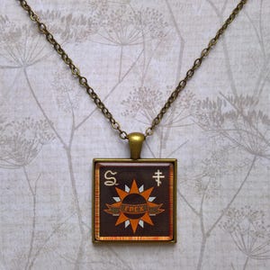 May include: Square pendant necklace with a bronze chain. The pendant has a dark brown background with an orange sunburst design and the word "ГРЕХ" on a banner. The pendant also features symbols and a bronze frame.