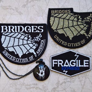 May include: Two black embroidered patches featuring a white and gold map of the United States with the text BRIDGES UNITED CITIES OF AMERICA, a black hexagonal patch with the text FRAGILE EXPRESS, and a black pendant necklace with a white handprint.