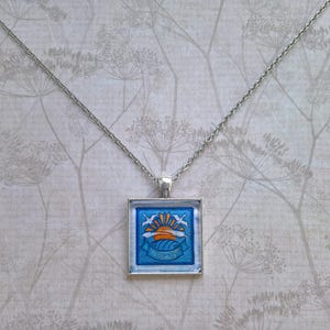 May include: Silver-toned necklace with a square pendant featuring a blue and orange sunrise design. The pendant has a silver frame and a chain. The design includes birds and waves, creating a nautical theme.