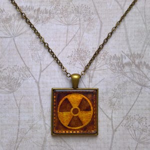 May include: A gold chain necklace with a square pendant featuring a yellow and brown radiation symbol.
