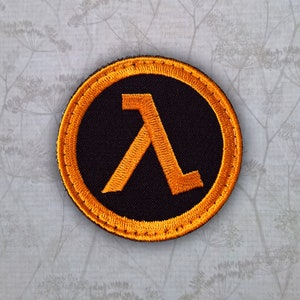 May include: A round, black embroidered patch with an orange Lambda symbol in the centre. The patch has a gold-coloured border.