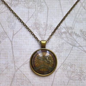 May include: A round pendant necklace with a bronze chain. The pendant features a detailed image of a bird with outstretched wings, set against a textured background with a banner below. The pendant has a bronze-colored frame.