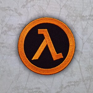 May include: A circular embroidered patch featuring a black background with a bright orange border and a centered orange Greek lambda symbol. The design is stitched with a dense thread texture, creating a raised appearance on the fabric surface.