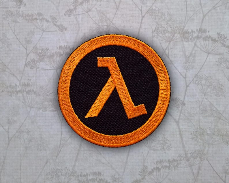 May include: A circular embroidered patch featuring a black background with a bright orange border and a centered orange Greek lambda symbol. The design is stitched with a dense thread texture, creating a raised appearance on the fabric surface.