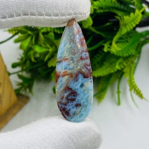 Può includere: Una gemma a forma di goccia, lucidata, con un motivo marmorizzato di tonalità azzurre, rosse e marroni. La pietra è tenuta su uno sfondo bianco con fogliame verde. La gemma è probabilmente destinata alla creazione di gioielli.