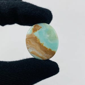 Puede incluir: Gema ovalada con superficie pulida. La piedra presenta una mezcla de colores, incluyendo azul claro, marrón y blanco, creando un efecto marmoleado. La gema se sostiene sobre un fondo negro.