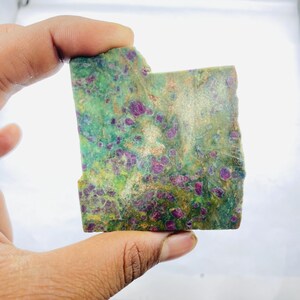 Puede incluir: Una losa rectangular de piedra natural con un color base verde y dorado, salpicada de numerosos puntos de color rojo intenso. La piedra tiene un borde áspero e irregular y una superficie texturizada, mostrando su estado bruto y sin pulir. La piedra se sostiene en una mano.