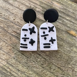 Puede incluir: Un par de pendientes hechos a mano. Cada pendiente presenta un perno circular negro conectado a un colgante blanco moteado en forma de arco. Los colgantes están decorados con formas geométricas negras. Los pendientes se exhiben sobre una superficie de madera.