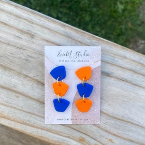 Puede incluir: Un par de pendientes hechos a mano con tres formas geométricas apiladas en azul y naranja. Los pendientes están en una tarjeta blanca con el texto "Evie M Studio Jacksonville, Alabama" y "Handcrafted in the USA".