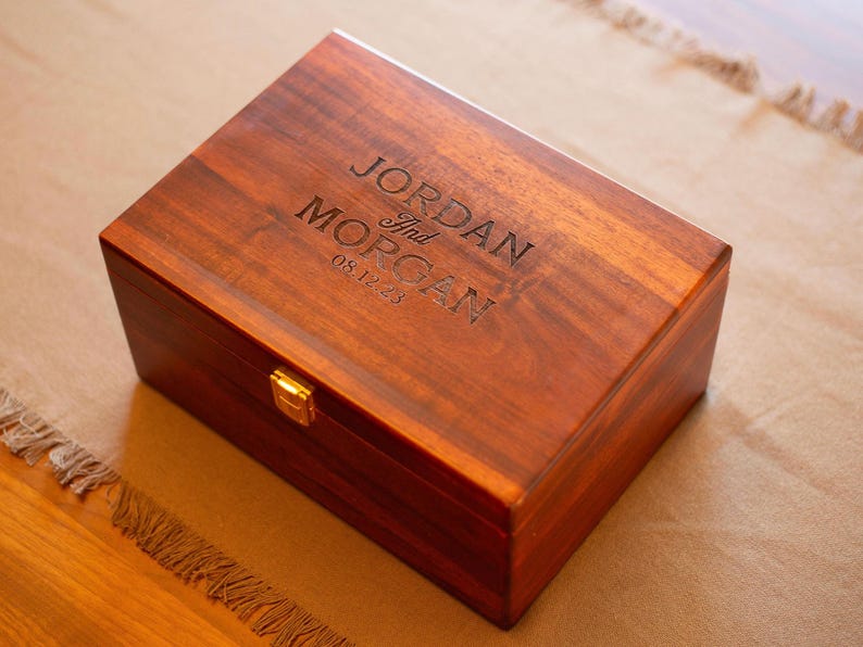 May include: A wooden box with a hinged lid and a brass latch. The box is engraved with the text "Jordan and Morgan 08.12.23".