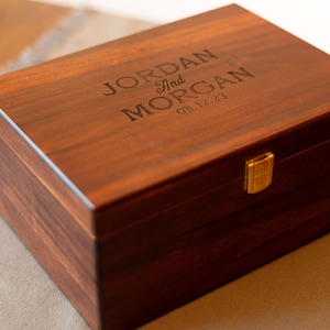May include: A dark brown wooden box with a gold latch. The top of the box is engraved with the names "JORDAN and MORGAN" and the date "08.12.23".