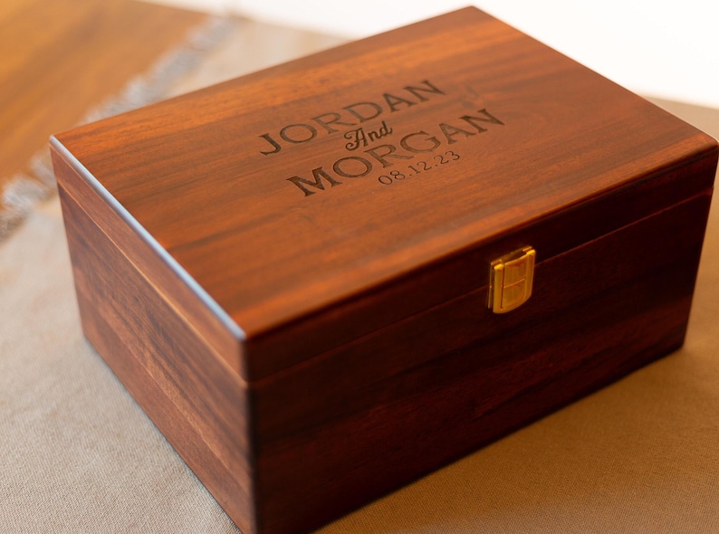 May include: A dark brown wooden box with a gold latch. The top of the box is engraved with the names "JORDAN and MORGAN" and the date "08.12.23".