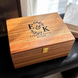 May include: A wooden box with a hinged lid and a gold latch. The box is engraved with the initials "E & K", the names "Ethan & Katie", and the date "8/2/2025".