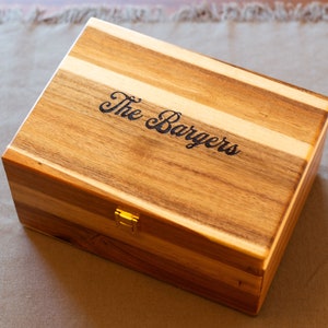 May include: A wooden box with a hinged lid and a brass latch. The box is made of light and dark wood and has a personalized inscription that reads "The Bargers".